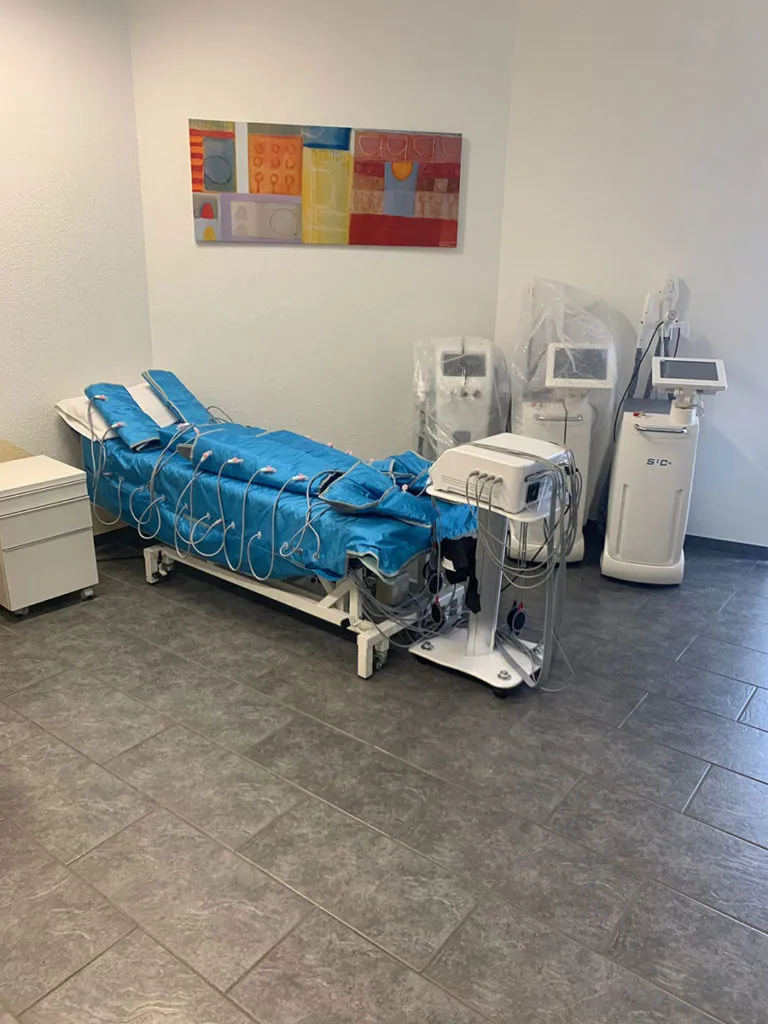 Behandlungsraum mit Liege und Pressotherapie-Gerät sowie weiteren Geräten
